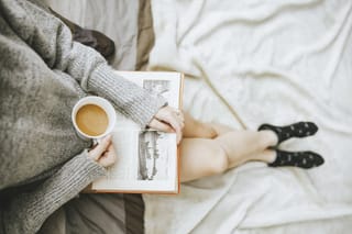 Person relaxes in bed with an open book and a warm cup of coffee