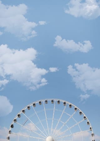 Top of a Ferris wheel rises into a bright blue sky with scattered clouds