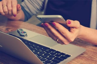 Hands use a smartphone beside an open laptop on a work table