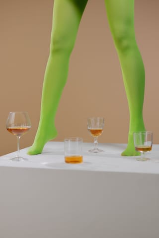 Legs in vivid green tights stand on a tabletop beside glasses and small objects