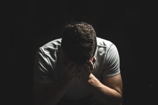 Man sits with his head lowered into his hands against a dark studio background
