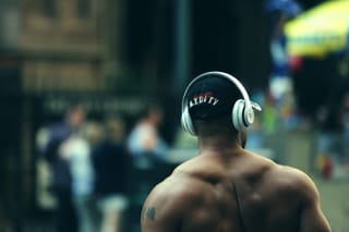 Muscular man wearing over-ear headphones is photographed from behind in a gym setting