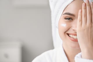 Smiling woman with a towel wrapped around her head applies facial skincare cream