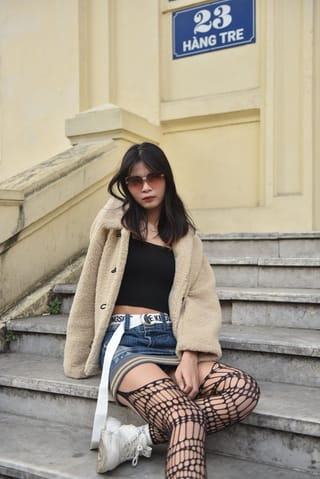 Woman sits on outdoor steps in a beige coat, denim shorts, and patterned tights