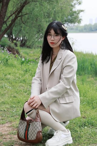 Woman in a light beige suit crouches outdoors beside greenery and water
