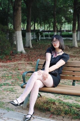 Woman sits on a park bench in a black dress, white tights, and heels