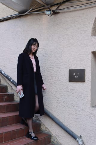 Woman in a long black coat stands beside outdoor stairs holding a small bag