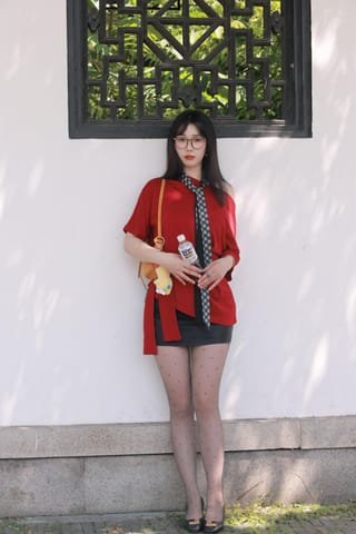 Woman stands beside a light wall wearing a red top and black skirt