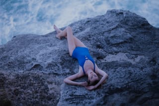 Woman in a blue outfit lies across large coastal rocks beside the sea