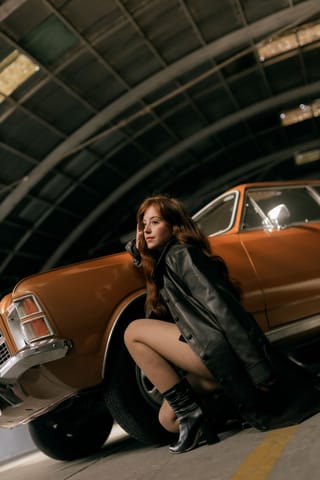 Woman wearing a black coat beside a classic car in a garage.