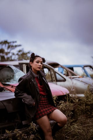 Woman in a plaid skirt posing beside abandoned cars.